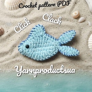 May include: A light blue crocheted fish applique with a dark blue stitched border and a small black bead eye. The text "Crochet pattern PDF", "Click Clack", and "Yarnproductsua" are visible. The fish is placed on a sandy surface with seashells.