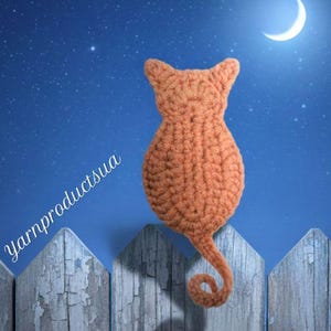 May include: An orange crochet cat silhouette sits atop a weathered wooden fence against a night sky backdrop with a crescent moon. The cat's tail curls, and the text "yarnproductsua" is visible on the left side of the image.