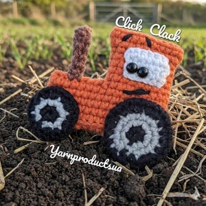 Puede incluir: Un juguete de tractor naranja tejido a ganchillo con ruedas negras y grises, una chimenea marrón y una cara blanca con ojos negros. El texto "Click Clack" está encima del tractor, y "Yarnproductsua" debajo.