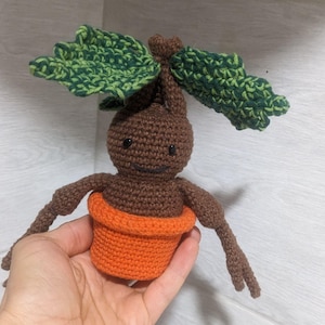 May include: A brown crocheted plant creature with green leaves and a smiling face, sitting in an orange crocheted pot.