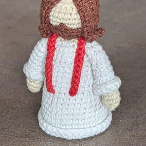 Może przedstawiać: Ręcznie robiona szydełkowa lalka przedstawiająca postać religijną. Lalka ma białą szatę z czerwonymi paskami, brązową brodę i włosy oraz jasnobeżową twarz. Lalka ma około 13 cm wysokości.