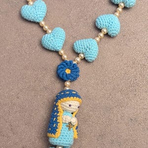 Puede incluir: Un rosario de ganchillo hecho a mano con cuentas en forma de corazón de color azul claro intercaladas con pequeñas perlas de color crema. El rosario incluye una cuenta en forma de flor azul oscuro, una figura de la Virgen María y una cruz de cuatro pétalos.