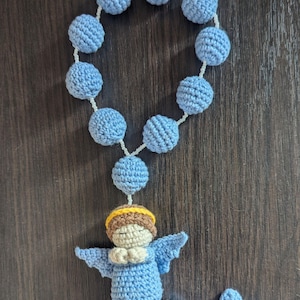 Puede incluir: Un rosario hecho a mano con ganchillo, con cuentas azul claro y una figura de ángel. El ángel tiene las manos juntas, alas y un halo. Una cruz de ganchillo está al final. Sobre un fondo de madera oscura.