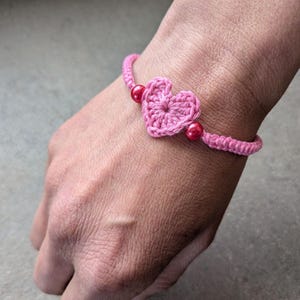 May include: Pink crochet bracelet featuring a heart-shaped centerpiece and two red beads. The bracelet is handmade with a delicate, textured design. The bracelet is a charming accessory.