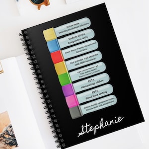 May include: A black spiral-bound notebook with colorful tabs and the name "Stephanie" in white script. The tabs list medical tests, including "EDTA Hematology, HbA1c" and "Blood bank testing." Sunglasses and a gold binder clip are also visible.
