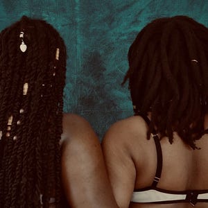 Op de afbeelding: Twee mensen met donker haar staan ​​naast elkaar, weg van de camera. De ene heeft lang, gevlochten haar versierd met gouden en zilveren accenten, waaronder een hartvormige bedel. De andere heeft korte dreadlocks. Beide dragen beha's.