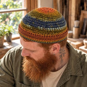 May include: A crochet beanie featuring a horizontal striped pattern in shades of orange, olive green, and navy blue. The hat has a rounded crown and a folded brim, constructed with a textured stitch pattern throughout the entire piece.