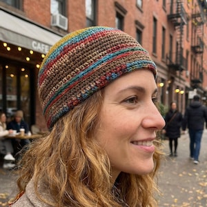 May include: A crocheted beanie featuring horizontal stripes in shades of brown, blue, red, and yellow. The hat has a textured, dense stitch pattern and fits snugly around the head, showcasing a multi-colored yarn design with earthy tones.