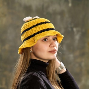 Può includere: Una donna indossa un cappello all'uncinetto a righe gialle e nere con un motivo a forma di ape. Il cappello ha dei pon pon bianchi in cima. Indossa un maglione nero a collo alto.