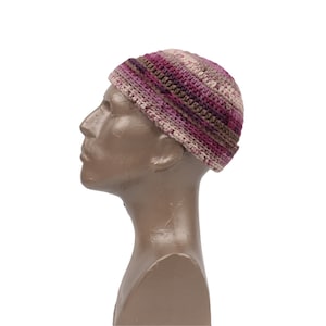 May include: A crocheted beanie hat with a striped pattern in shades of pink, brown, and beige. The hat is shown on a mannequin head.