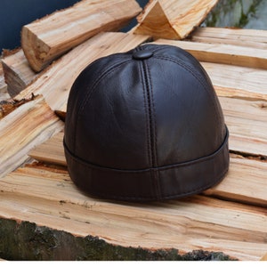 May include: A dark brown leather cap with a rounded crown and a small button on top. The cap has a short brim and is made of smooth leather. The cap is sitting on a pile of wood.
