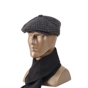 May include: A gray and black houndstooth pattern newsboy cap. The cap is displayed on a mannequin head with a black scarf. The cap has a rounded crown and a small, stiff brim. The scarf is draped around the mannequin's neck.