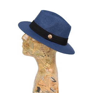 May include: A blue and black fedora hat with a gold button on the front. The hat is made of a woven material and has a wide brim.