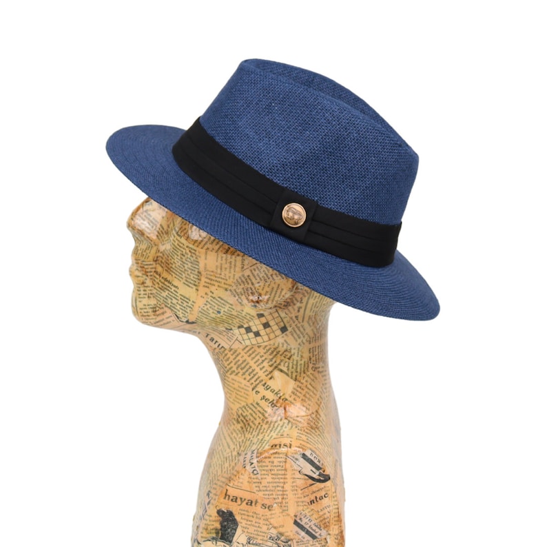 May include: A blue and black fedora hat with a gold button on the front. The hat is made of a woven material and has a wide brim.