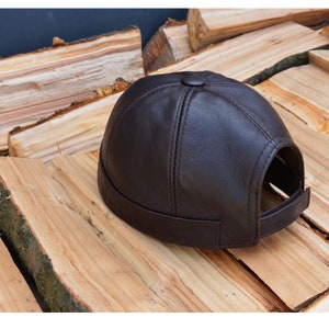 Puede incluir: Una gorra de béisbol de cuero marrón con una corona redonda y una visera plana. La gorra tiene una costura cosida en el centro de la corona y un pequeño botón en la parte superior.