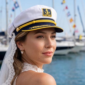 May include: A white captain's hat with a navy blue brim and gold accents. The hat features an anchor emblem and the word "CAPTAIN". The hat is worn with a white lace veil and pearl earrings. The background includes sailboats and water.