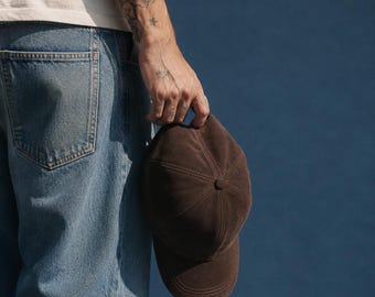 Könnte beinhalten: Eine dunkelbraune Baseballkappe mit glatter, matter Textur. Die Kappe verfügt über eine klassische Sechs-Panel-Konstruktion mit sichtbaren, farblich abgestimmten Nähten entlang der Segmente und einer abgerundeten Krone. Der Schirm ist gebogen.