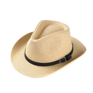 Może przedstawiać: Beżowy kapelusz typu fedora z czarnym skórzanym paskiem i klamrą. Kapelusz ma szerokie rondo i strukturalną koronę. Kapelusz jest wykonany z tkanego materiału, prawdopodobnie słomy lub podobnej tkaniny.