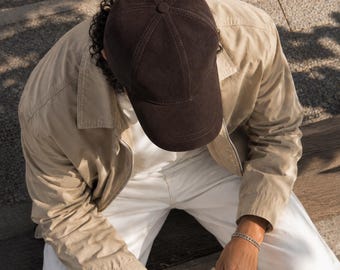 Könnte beinhalten: Eine dunkelbraune Baseballkappe aus Wildleder mit sechs Paneelen und einem Knopf oben, getragen zu einer beigen Jacke mit Kragen, einem weißen Hemd, einer weißen Hose und einem silbernen Kettenarmband, während ein Kaffeebecher gehalten wird.