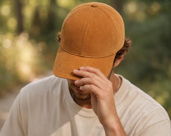 Könnte beinhalten: Eine einfarbige, hellbraune Baseballkappe mit glatter, matter Textur. Die Kappe verfügt über eine klassische Sechs-Panel-Konstruktion mit gebogenem Schirm und einem Knopf an der Oberseite. Das Material wirkt weich und erinnert an Wildleder.