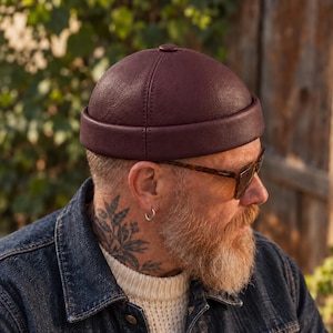 Puede incluir: Un gorro tipo docker de color burdeos oscuro con una textura suave similar al cuero, una corona redondeada, una costura central cosida y un borde grueso doblado que se asienta sobre las orejas, ofreciendo una silueta estructurada.