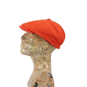 Könnte beinhalten: Eine leuchtend orangefarbene Flatcap mit leicht gebogenem Schirm. Die Kappe ist aus einem weichen, leichten Stoff gefertigt und hat ein klassisches, zeitloses Design.