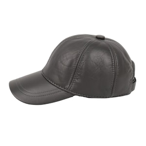 Puede incluir: Gorra de béisbol de cuero gris con visera curva y botón en la parte superior. La gorra presenta un diseño clásico de seis paneles con costuras y una correa ajustable en la parte trasera.