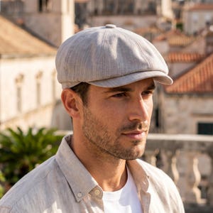 Może przedstawiać: Jasnoszara czapka typu newsboy z krótkim daszkiem. Czapka ma teksturowany, prążkowany wzór i guzik na górze. Osoba nosi jasną koszulę zapinaną na guziki i biały t-shirt.