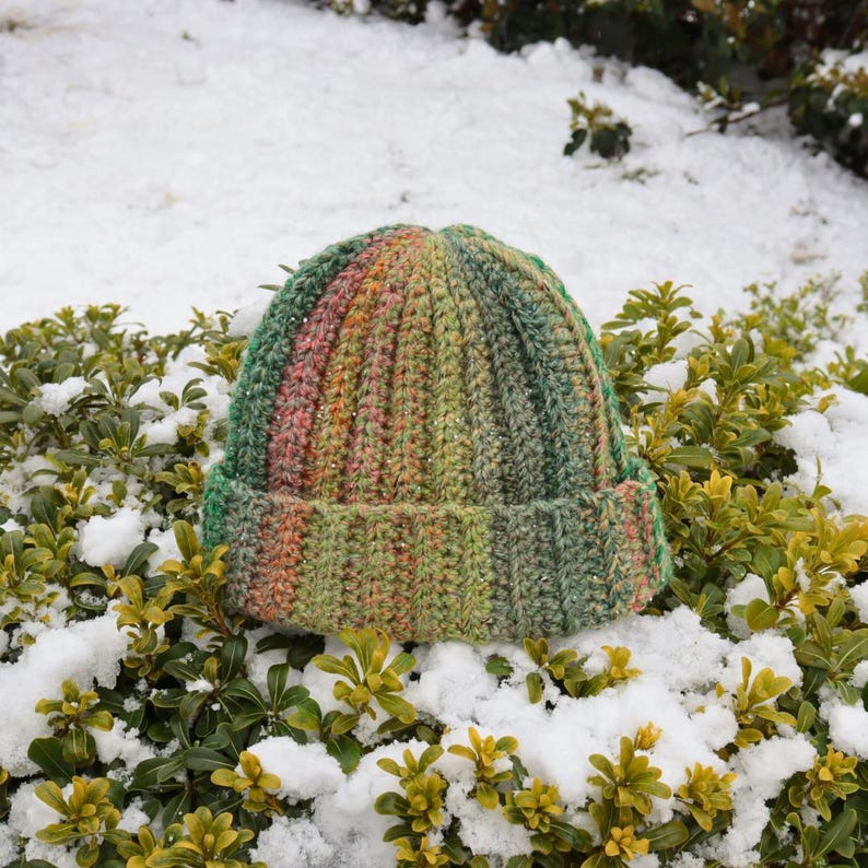May include: A handmade, ribbed knit beanie hat in shades of green, orange, and pink. The hat is folded at the brim and sits atop snow-covered greenery. The hat is a winter accessory.
