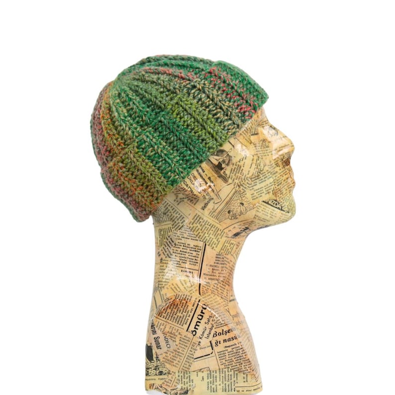 May include: A green and pink knit beanie hat with a ribbed design. The hat is displayed on a mannequin head covered in newspaper print. The beanie is a mix of green, pink, and yellow colors.