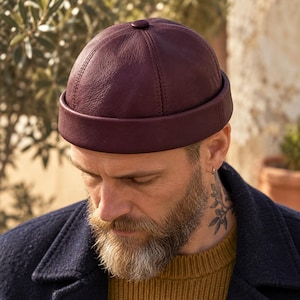 May include: A deep burgundy leather docker cap featuring a rounded crown with six stitched panels, a small central button at the top, and a wide, folded brim that encircles the head. The material has a visible grain texture.