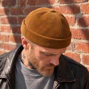 May include: A tobacco brown suede docker cap featuring a six-panel construction with visible stitching, a rounded crown, and a thick, folded brim. The material has a soft, matte texture, and the design is brimless, sitting close to the head.