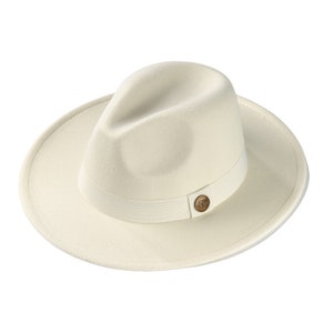 Könnte beinhalten: Ein cremefarbener Fedora-Hut mit breiter Krempe und eingedrückter Krone. Der Hut verfügt über ein passendes cremefarbenes Ripsband, das die Basis der Krone umschließt und mit einem kleinen, runden, bronzefarbenen Zierknopf versehen ist.