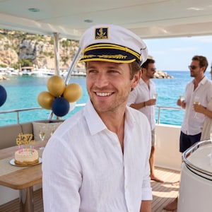 May include: A man wearing a white captain's hat with a gold anchor emblem and navy blue accents. He is smiling on a yacht, with a birthday cake and balloons in the background. Other people are visible, holding champagne glasses.