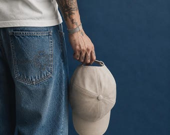 Könnte beinhalten: Eine einfarbige, beige Baseballkappe mit sechs-teiliger Konstruktion, einem zentralen Knopf auf der Krone und einem gebogenen Schirm. Die Kappe verfügt über einen Stoffriemen mit Metallschnallenverschluss auf der Rückseite für ein schlichtes Design.