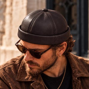 May include: A dark brown leather docker cap featuring a rounded crown with six stitched panels, a small top button, and a thick, folded brim. The cap has a textured surface and a structured fit, designed to sit above the ears.