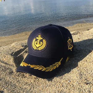 Op de afbeelding: Marineblauwe baseballcap met goud geborduurde nautische emblemen, waaronder een anker, een lauwerkrans en een scheepsstuur. De rand heeft een goud geborduurd bladerontwerp. De pet ligt op zand bij het water.