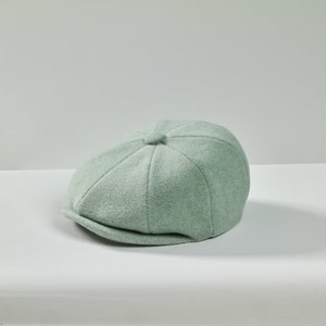 Peut inclure: Une casquette de newsboy en laine vert clair avec une couronne arrondie et une visière courte.