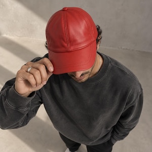 May include: A vibrant red baseball cap constructed from a smooth, leather-like material. The cap features a classic six-panel design with visible stitching along the seams and a curved brim. The top of the cap is finished with a small, matching red button.
