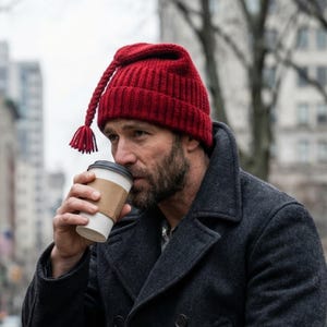 May include: A red knit hat with a long braided tassel and a matching tassel at the end. The hat is worn by a person wearing a dark gray coat. The person is holding a coffee cup.