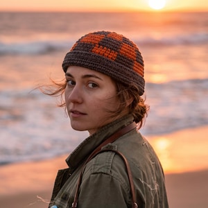 Puede incluir: Un gorro con vuelta que presenta un patrón de cuadros en gris oscuro y naranja brillante. El gorro está confeccionado con hilo de ganchillo y tiene un borde grueso y doblado que se ajusta a la cabeza, mostrando un diseño de punto texturizado.