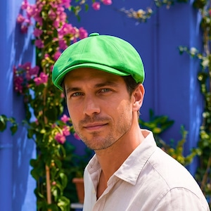 Könnte beinhalten: Eine leuchtend grüne Flatcap. Die Kappe besteht aus einem glatten Stoff und hat eine abgerundete Oberseite. Im Hintergrund eine blaue Wand mit rosa Blüten und Grün.