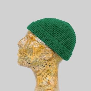 May include: A green crocheted beanie hat on a mannequin head. The hat is a simple design with a rounded crown and a folded brim.