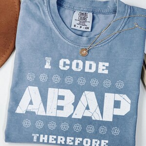 May include: A blue jean Comfort Colors t-shirt with the text "I CODE ABAP THEREFORE" in white. The shirt is folded and has a gold necklace draped across it. A brown hat and dried plant are in the background.