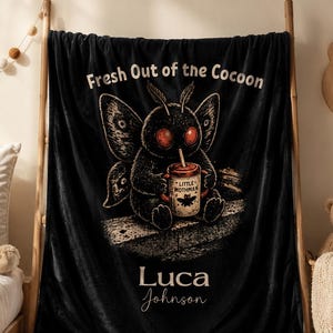 May include: Black plush blanket with the text "Fresh Out of the Cocoon" and a cartoon moth holding a coffee cup. The cup reads "Little Mothman". The name "Luca Johnson" is written at the bottom. The blanket is displayed on a wooden ladder.
