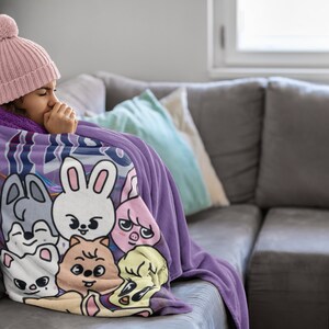 Puede incluir: Una persona con un gorro de punto rosa est&aacute; envuelta en una manta de forro polar morada con un estampado de animales de dibujos animados coloridos. La manta presenta una variedad de animales, incluyendo un conejo, un perro, un gato y un cerdo. La persona est&aacute; sentada en un sof&aacute; gris.