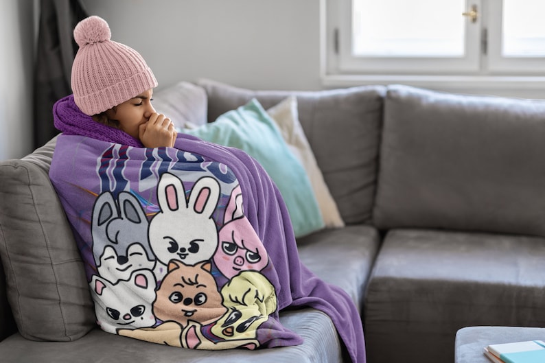 Puede incluir: Una persona con un gorro de punto rosa est&aacute; envuelta en una manta de forro polar morada con un estampado de animales de dibujos animados coloridos. La manta presenta una variedad de animales, incluyendo un conejo, un perro, un gato y un cerdo. La persona est&aacute; sentada en un sof&aacute; gris.