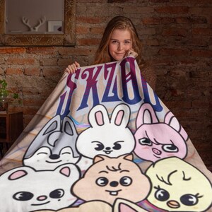 Puede incluir: Una manta de forro polar beige con un dise&ntilde;o de dibujos animados coloridos que presenta una variedad de animales, incluyendo un conejo, un cerdo, un p&aacute;jaro y un perro. La manta tiene el texto "SKZOO" impreso en azul y morado.