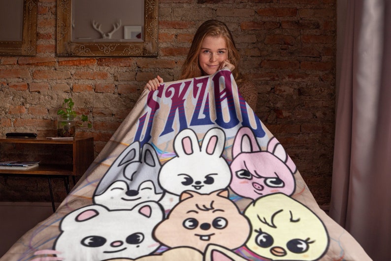 Puede incluir: Una manta de forro polar beige con un dise&ntilde;o de dibujos animados coloridos que presenta una variedad de animales, incluyendo un conejo, un cerdo, un p&aacute;jaro y un perro. La manta tiene el texto "SKZOO" impreso en azul y morado.