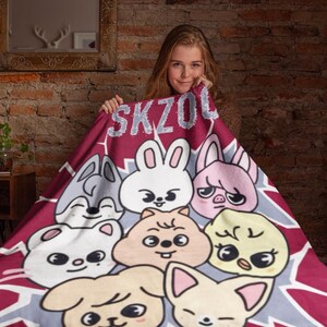 May include: A red and gray fleece blanket with a cartoon design featuring a variety of animals, including a rabbit, a pig, a wolf, a dog, and a bird. The blanket has the text "SKZOO" printed on it.